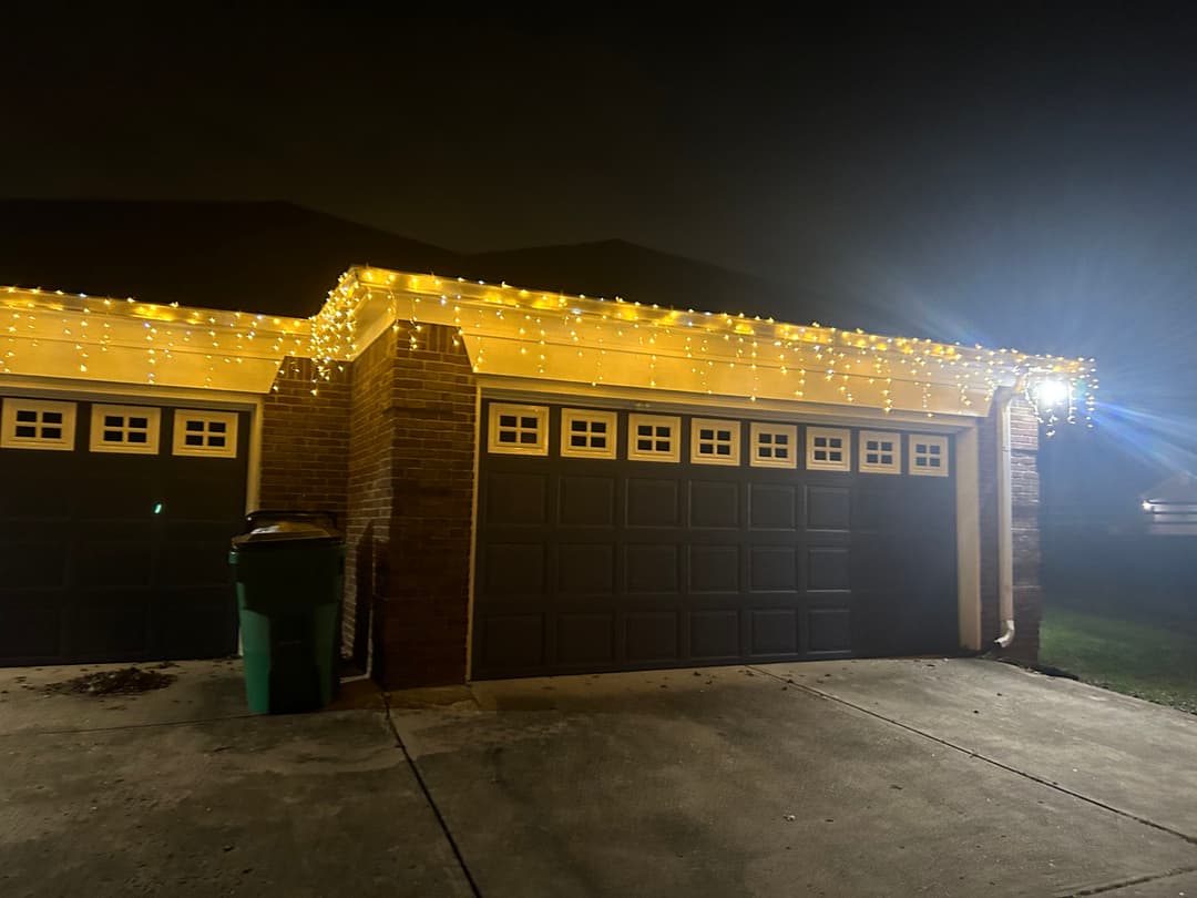 Garage adorned with yellow string lights at night, illuminated driveway and green trash bin visible.